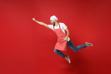 Crazy young male chef cook or baker man in striped apron white t-shirt toque chefs hat posing isolated on red background. Cooking food concept. Mock up copy space. Jumping, fooling around, having fun.