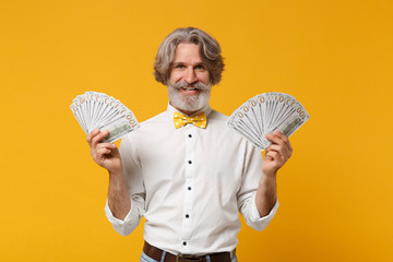 Smiling elderly gray-haired mustache bearded man in white shirt bow tie isolated on yellow orange background. People lifestyle concept. Mock up copy space. Hold fan of cash money in dollar banknotes.