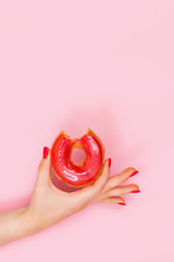 Hand of a beautiful woman holds a sweet red bitten donut with sparkles. Closeup of hands of a young woman with manicure on nails.