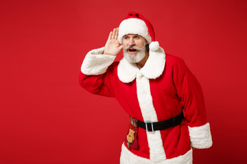 Fototapeta na wymiar Elderly gray-haired mustache bearded Santa man in Christmas hat posing isolated on red background. New Year 2020 celebration holiday concept. Mock up copy space. Try to hear you with hand near ear.