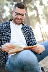 Fototapeta premium Hombre joven con lentes y camisa de cuadros apoyado en un árbol leyendo sonriente un libro al aire libre
