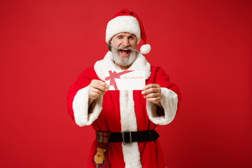 Cheerful elderly gray-haired mustache bearded Santa man in Christmas hat isolated on red wall background. Happy New Year 2020 celebration holiday concept. Mock up copy space. Holding gift certificate.