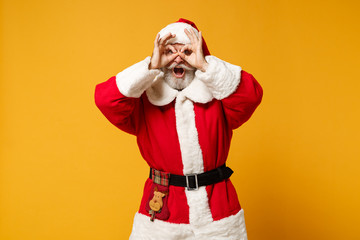 Fototapeta na wymiar Elderly gray-haired mustache bearded Santa man in Christmas hat isolated on yellow background. New Year 2020 celebration concept. Mock up copy space Hold hand near eye imitating binoculars or glasses.