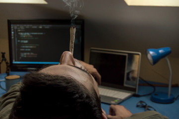 Young man smoking a marijuana joint at home while working with computers and programming code....