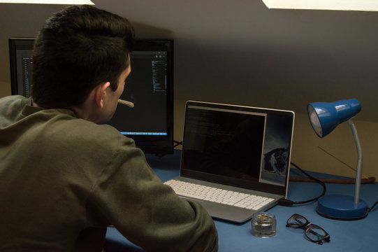 Concentrated Young Man Smoking A Marijuana Joint At Home While Working With Computers. Concept Of Cannabis And Working With Technology.