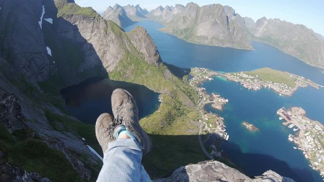 POV shot. Alone traveler sitting on a mountain edge above a Reine city, Lofoten, Norway. Close up of woman hiking boots.