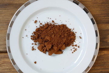 Brown vegetable carob (Ceratonia silica) for making a natural caffeine-free sweet drink. Dry carob powder in a white plate on a wooden background.