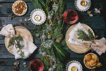 Christmas or New year table setting with empty ceramic plates, wine glasses, napkins, Christmas thuja wreath, luminous garland and burning candles on dark wooden plank table. Holiday mood flat lay