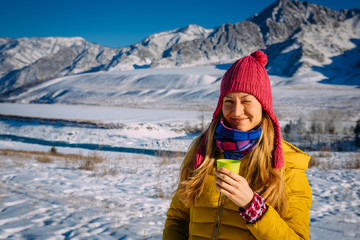 Fototapeta na wymiar Beautiful girl in red hat holds glass on the background of snow-capped mountains on frosty sunny day. Cheerful young woman enjoying Christmas vacation in the mountains, looking at camera and smiling.