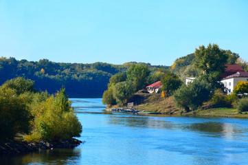 Obraz premium turn, bend of the river against the background of a darkening forest in the distance and blue sky. On the right you can see the buildings and a small pier on the water