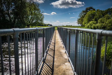 High contrast, shallow focus of a narrow footbridge seen crossing a large weir in the English countryside in early summer.