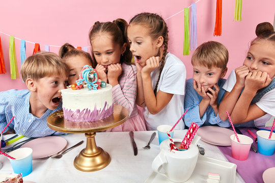 little girls and boys with wide open mouths loking at the cake with amazed expression. isolated pink background