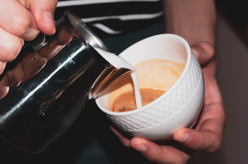 close up of barista hands pouring milk from metal jug into white cup with coffe in cafe