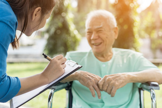 Female care worker serving checklist health to Happy smiling Asian senior man in a wheelchair relaxing while outside on park at Good environment and fresh air.Assistant,Retirement or elderly caregiver