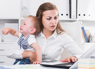 Female is working with a naughty child in her arms behind a laptop