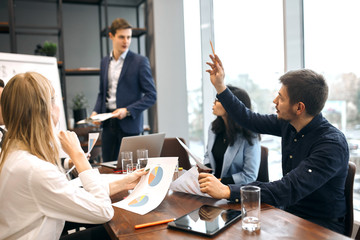 handsome clever man raising his arm, asking a question during the lecture, meeting, panorama window in the background of the photo. business people discussing the project, chart. Can I askyou question