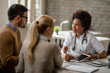Smiling black female doctor using touchpad while talking to a couple at clinic.