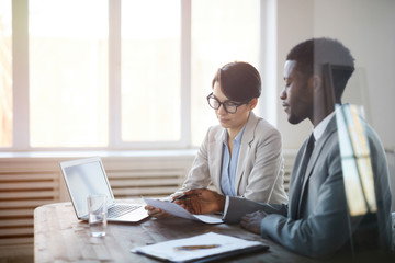 Obraz premium Portrait of two young entrepreneurs discussing business project while sitting at meeting table behind glass wall, copy space