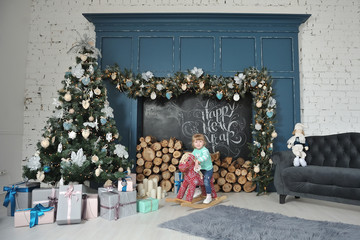 Little girl child sitting on a red rocking horse and smiling on the background of the Christmas interior with a fireplace, firewood, gifts and a Christmas tree