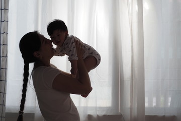 Silhouette tone of Asian mother raising her cute baby up and playing with him at home with white curtains background. Happiness of being motherhood, lovely warm moment mom and child, lifestyle concept