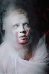 halloween make up. scary man ready for party. guy with face covered in white paint and bloody handprint mark on black studio background with fog