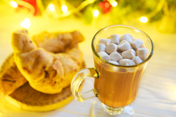 Winter hot chocolate with marshmallows and a croissant on a white wooden table against the background of the Christmas tree