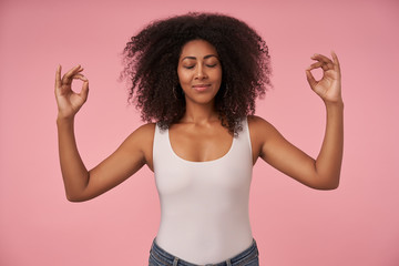 Pleasant looking young curly female with dark skin keeping eyes closed while meditating, posing...