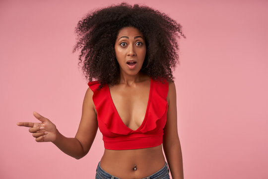 Young bemused curly dark skinned woman with belly button piercing pointing aside with forefinger while posing over pink background, looking to camera with wde eyes and mouth opened