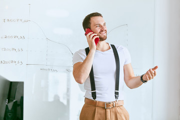 Young businessman wearing white t-shirt and trousers with suspenders look side, talking on phone with business partners. Flipchart diagrams background