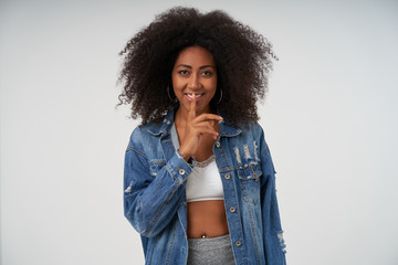 Positive young dark skinned lady with curly hair wearing casual clothes over white background, looking at camera with cheerful smile and holding index finger on her lips, asking to keep secret