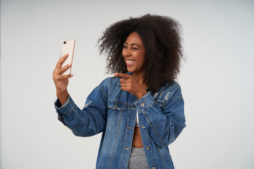 Indoor portrait of cheerful young curly dark skinned woman in casual clothes holding smartphone in raised hand, having pleasant talk with video chat, smiling happily over white background