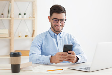 Young male in trendy glasses exchanging messages on smartphone while working in office