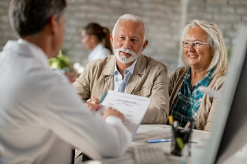 Happy senior couple going through medical insurance paperwork with a doctor. - obrazy, fototapety, plakaty