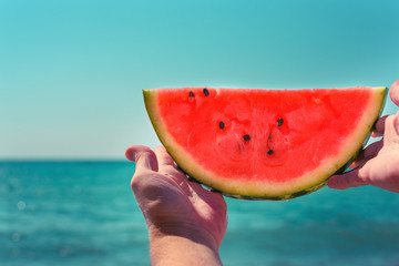 Watermelon, juicy, red slice in the hands of woman against the background of the ocean, in the rays of sunlight. Diet of tropical fruits.