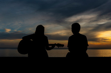 The silhouette of a woman playing guitar with a boyfriend at the seaside in the sunset.