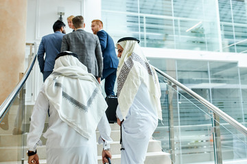 Two sheikhs go up stairs in business center. Foloowing caucasians in tuxedo, suits
