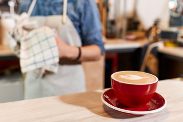 Beautiful big red cup of coffee stands on light coloured wooden table, heart cappuccino art, background of barista with towel, close up