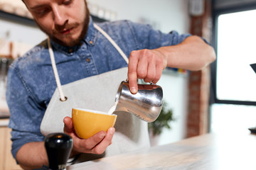 Confident concentrated bartender in neat apron pours steamed hot milk from metal jug into brightly coloured small coffee cup, work process concept