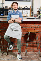 confident young barista wearing blue shirt, jeans and snickers, in white apron, sits on chair with hands crossed over, politely looks straightly at camera, expresses friendliness, indoor shot