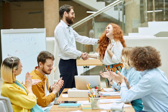 Pleased smart woman shakes hand with formally dressed man, keeps hand on heart with thankfull expression, people enthusiastically clap hands in brightly lighted office