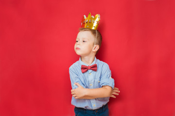 Emotional education and childhood concepts with an adorable 6-year old boy holding a golden king crown on his head as a wise spoiled child, red background