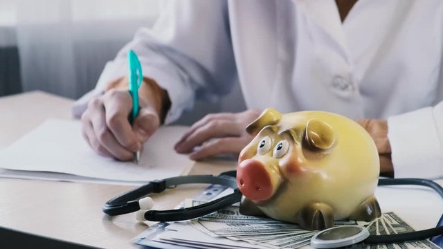 The doctor writes something on the background of a piggy bank and dollars lying on the table. The concept of the prestige of the work of a doctor, medical insurance and pharmacology, saving money on a