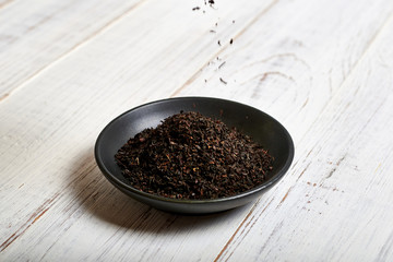 Black dry tea in a clay bowl on a light wooden background.