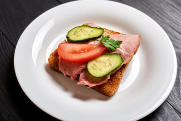 Sandwich with meat and vegetables on a white plate on a black wooden background.