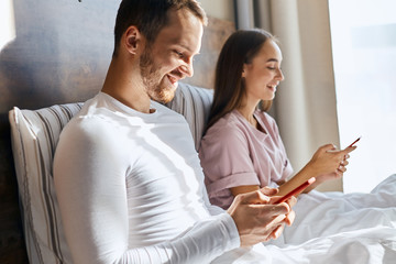 Pleasant young wife and husband chatting with parents using cell phones, getting good news, smiling broadly, looking aside with happy faces, in bedroom, side shot