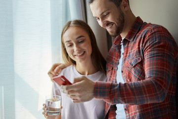 Charming lovely young couple standing near bright window, handsome smiling man hugs pretty wife, shows funny video on cell phone, having positive time together at home