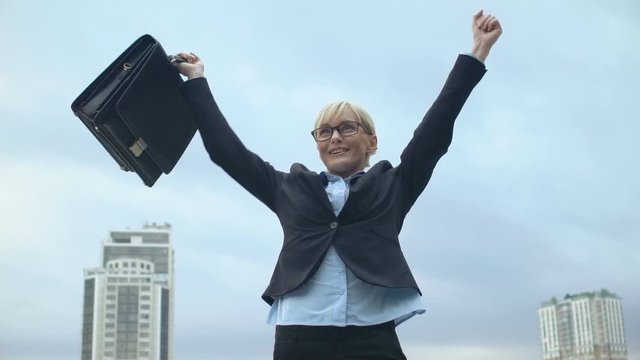 Happy woman in office suit holding bag and raising hands in victory gesture