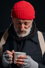 Tired and sick homeless male in coat and red hat look with sadness at mug. Portrait of bearded man...