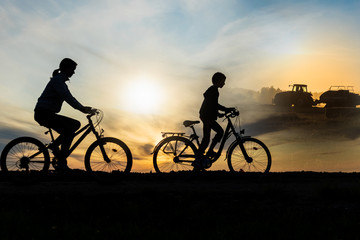 Boy , kid 10 years old, and girl riding bikes in countryside, tractor working in background,  silhouette of riding persons and machine at sunset in nature