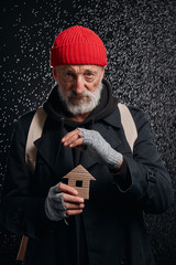 Senior street man wearing coat and red hat, holding house made by cardboard dream about own shelter